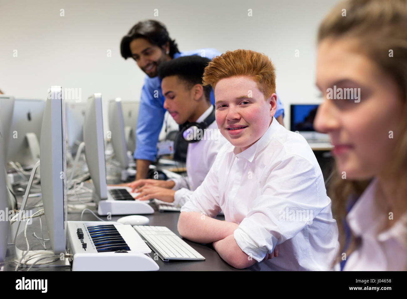 Students using computers in a school lesson. One student is smiling at the camera. There is a teacher helping another student in the background. Stock Photo