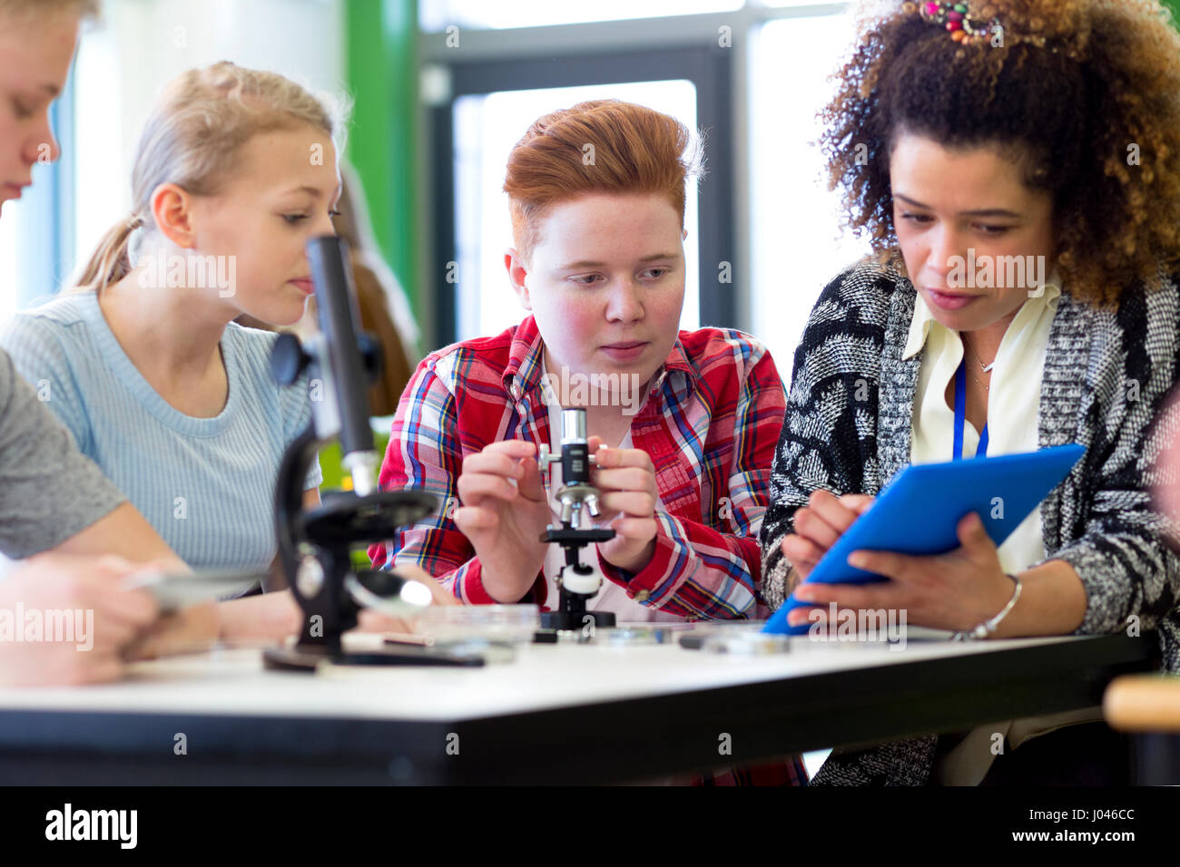 Students sitting with teacher in a lesson. They are using microscopes and a digital tablet. Stock Photo