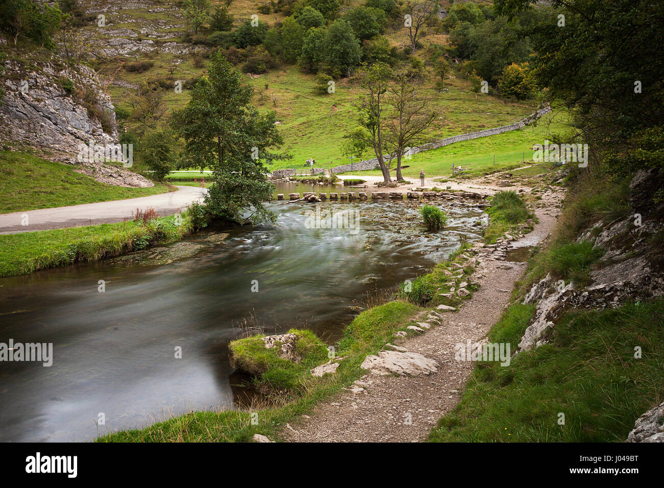 A view toward the stepping stones on the river Dove in Dovedale, Derbyshire. Slow shuuter speed blurs the water and several blury shaddows of people a Stock Photo