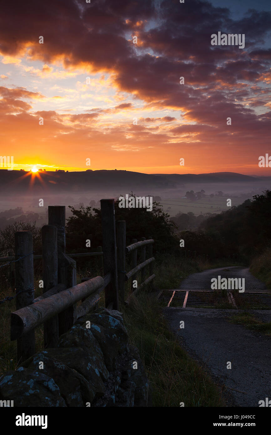 A colourful sunrise above the village of Longnor in North Staffordshire. A farm track is seen heading toward the sunrise with a wooden fence. Stock Photo