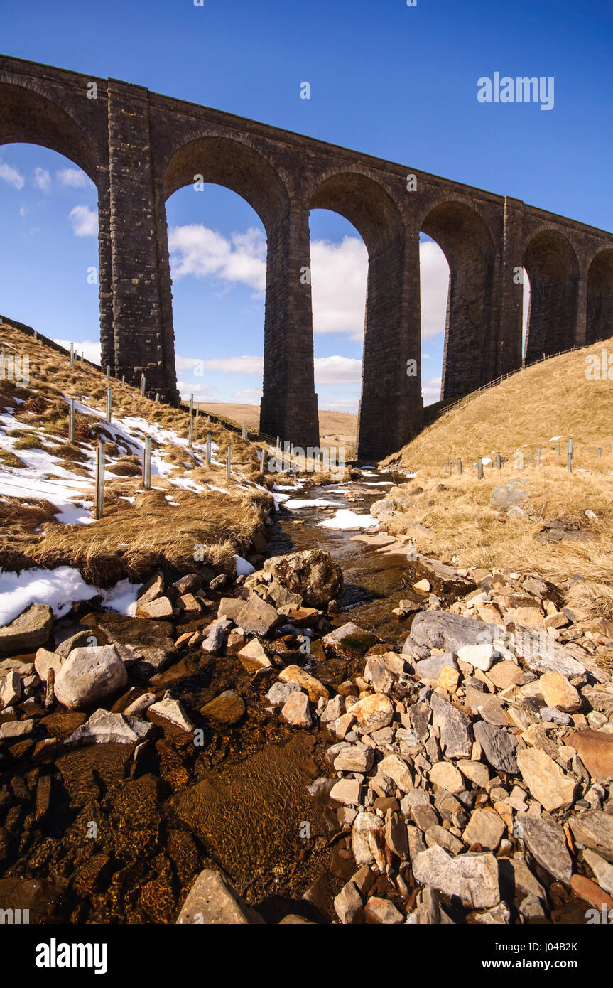 The Arten Gill Viaduct on the Settle to Carlisle Railway Line is silhouetted against a blue sky above the snow-speckled mountain stream of Arten Gill  Stock Photo