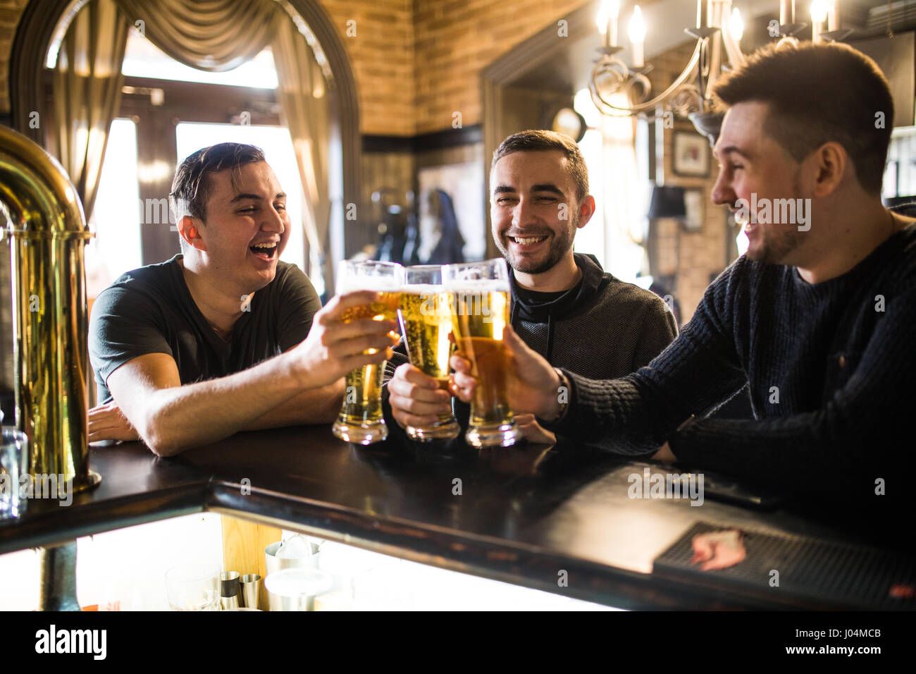 Three happy young men in casual wear talking and drinking beer while sitting at the bar counter together Stock Photo