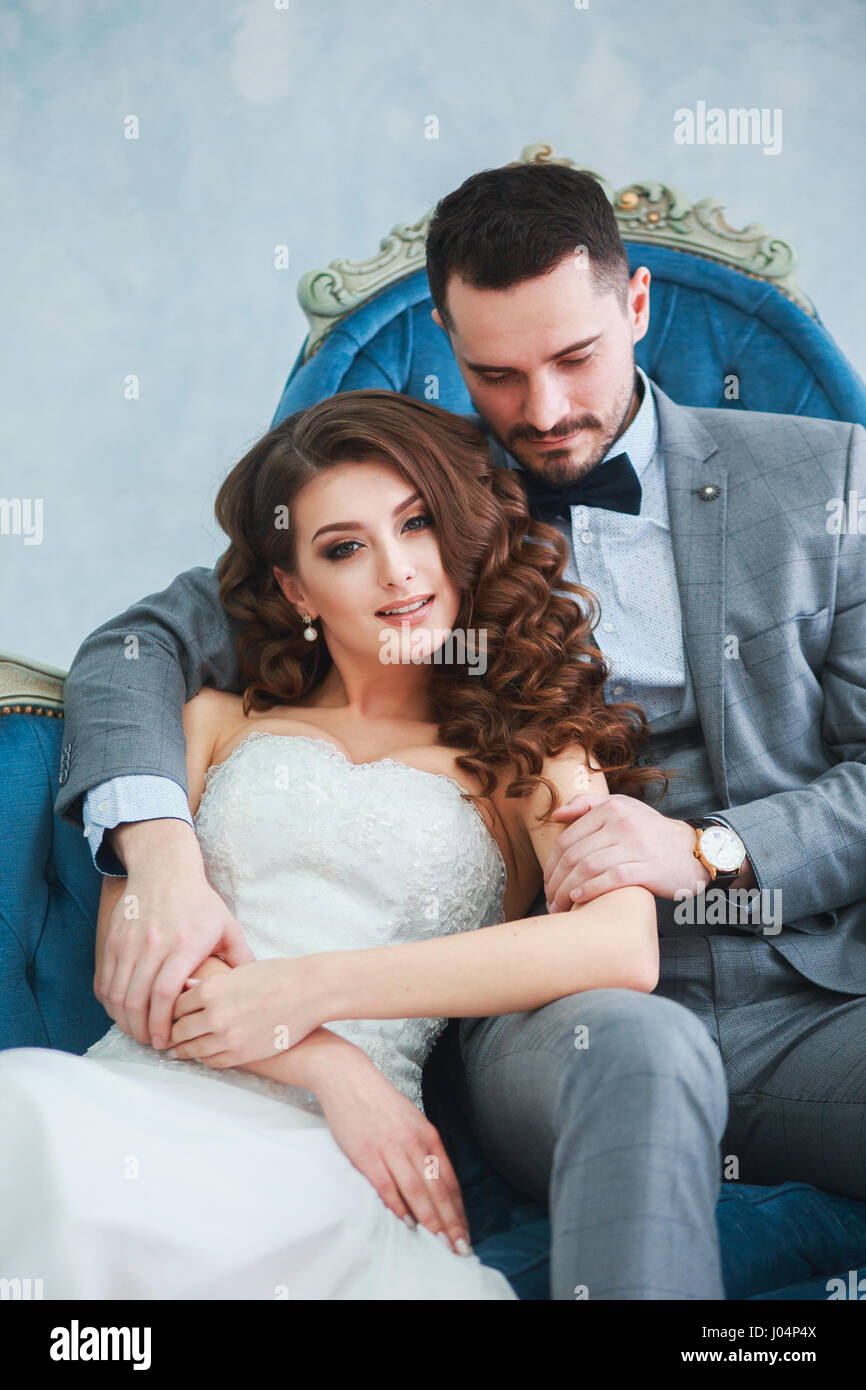 Bride in beautiful dress and groom in gray suit sitting on sofa indoors ...