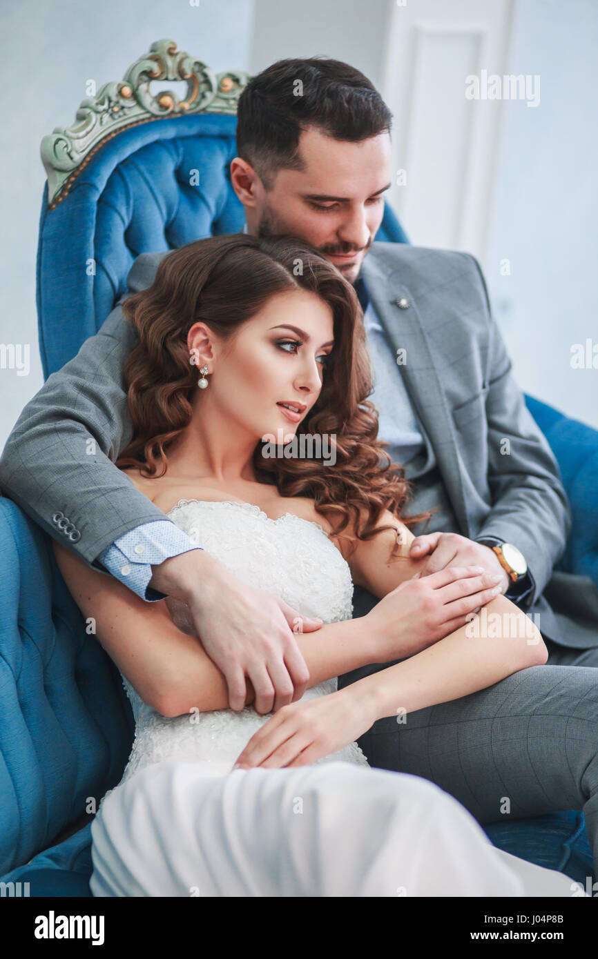 Bride in beautiful dress and groom in gray suit sitting on sofa indoors ...