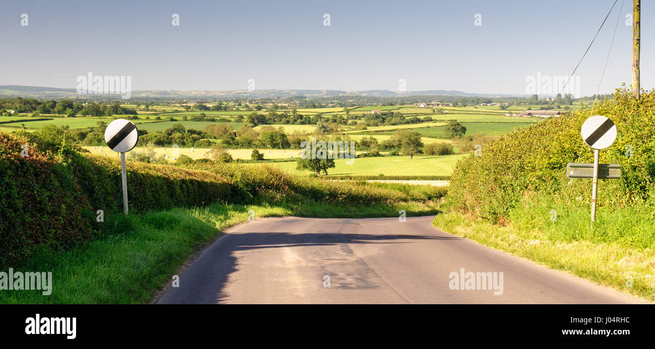 A narrow country lane with national speed limit signs descends a hill through dairy pasture fields at Cox Hill in Marnhull, in the rolling landscape o Stock Photo