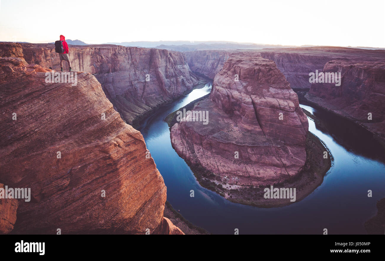 A male hiker is standing on steep cliffs enjoying the beautiful view of Colorado river flowing at famous Horseshoe Bend overlook in twilight, Arizona Stock Photo