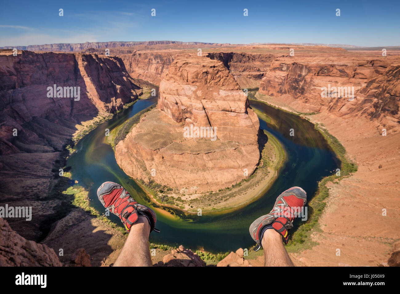 Wide-angle view of a young hiker hanging his feet off a cliff at famous Horseshoe Bend on a beautiful sunny day with blue sky and clouds, Arizona, USA Stock Photo