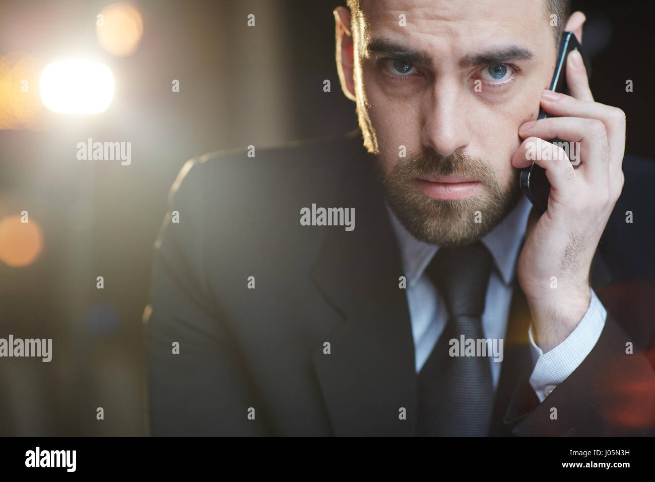 Portrait of modern businessman wearing black formal suit looking at camera while busy talking by mobile phone against black background with lens flare Stock Photo