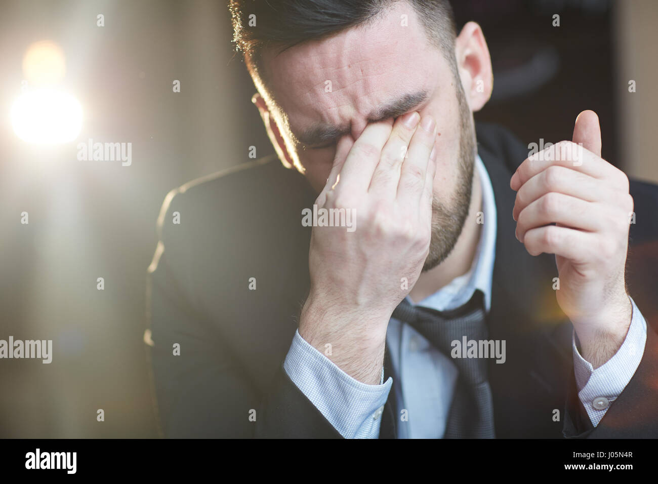 Portrait of tired adult man rubbing eyes and crying against black background with lens flare Stock Photo