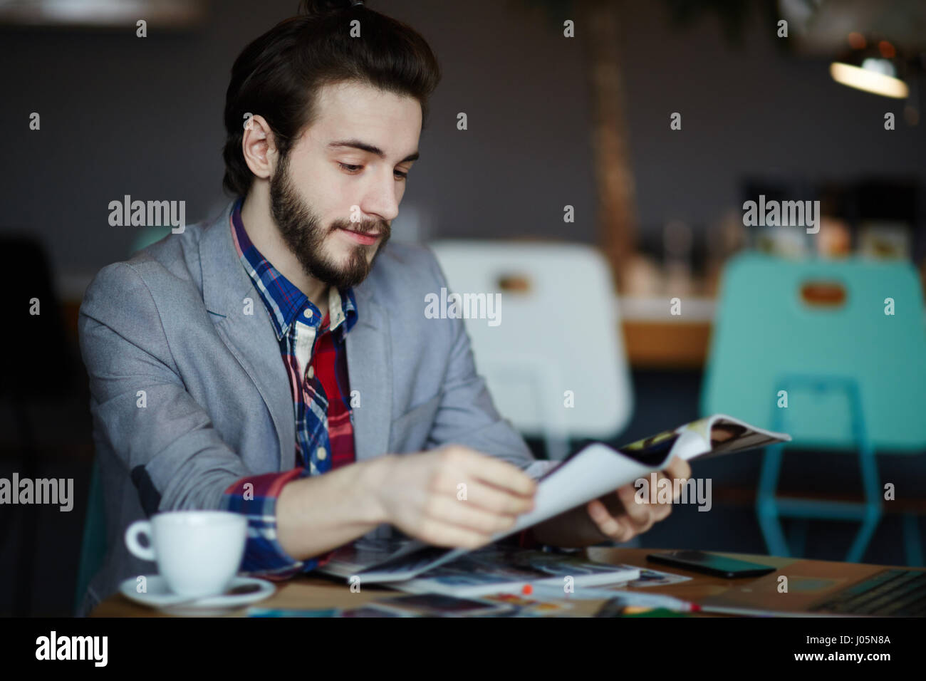 Portrait of confident long haired man wearing business casual clothes sitting at table in cafe reading magazines Stock Photo