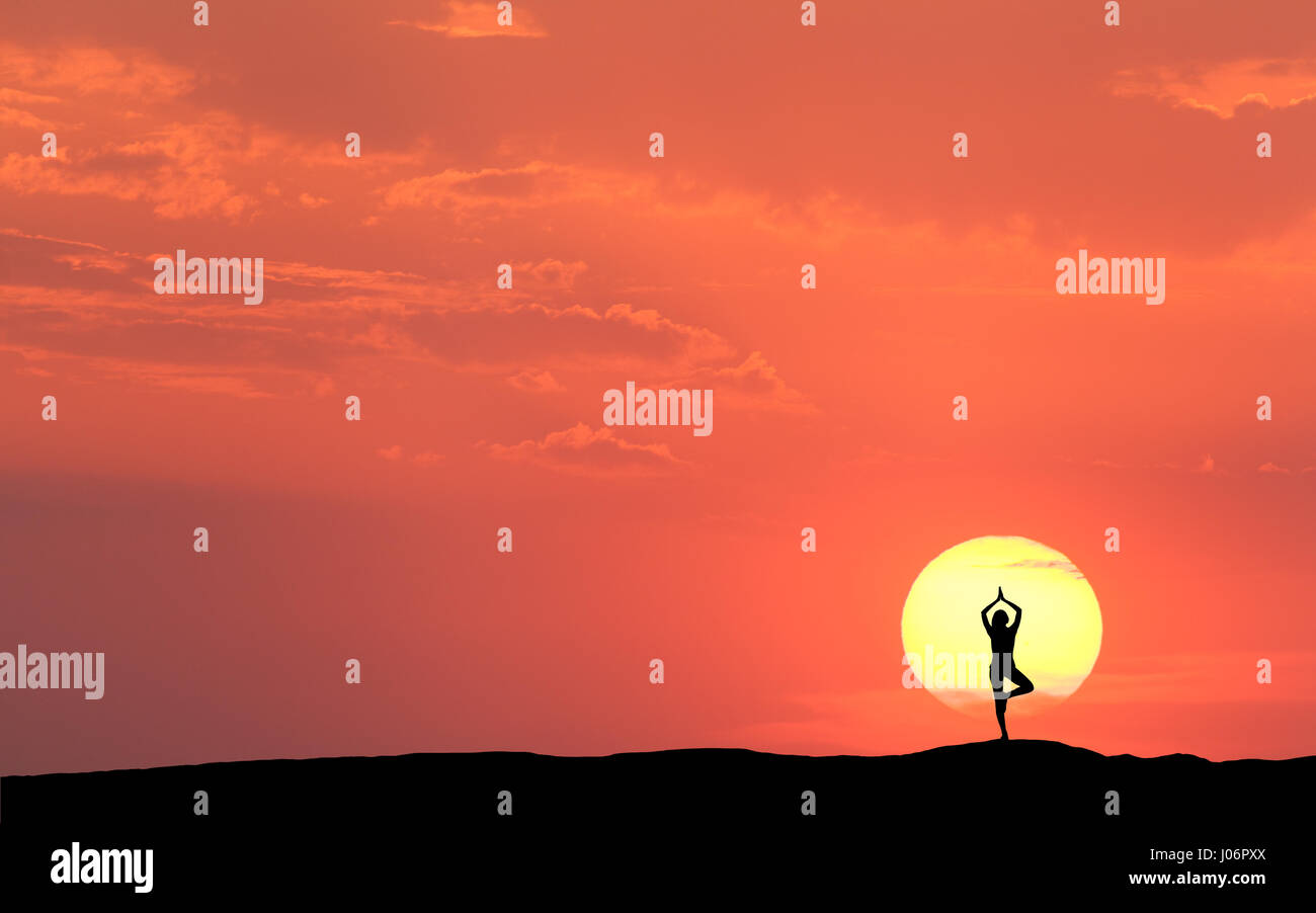 Silhouette of a standing sporty woman practicing yoga with raised up arms on the hill on the background of sun and colorful orange sky with clouds Stock Photo