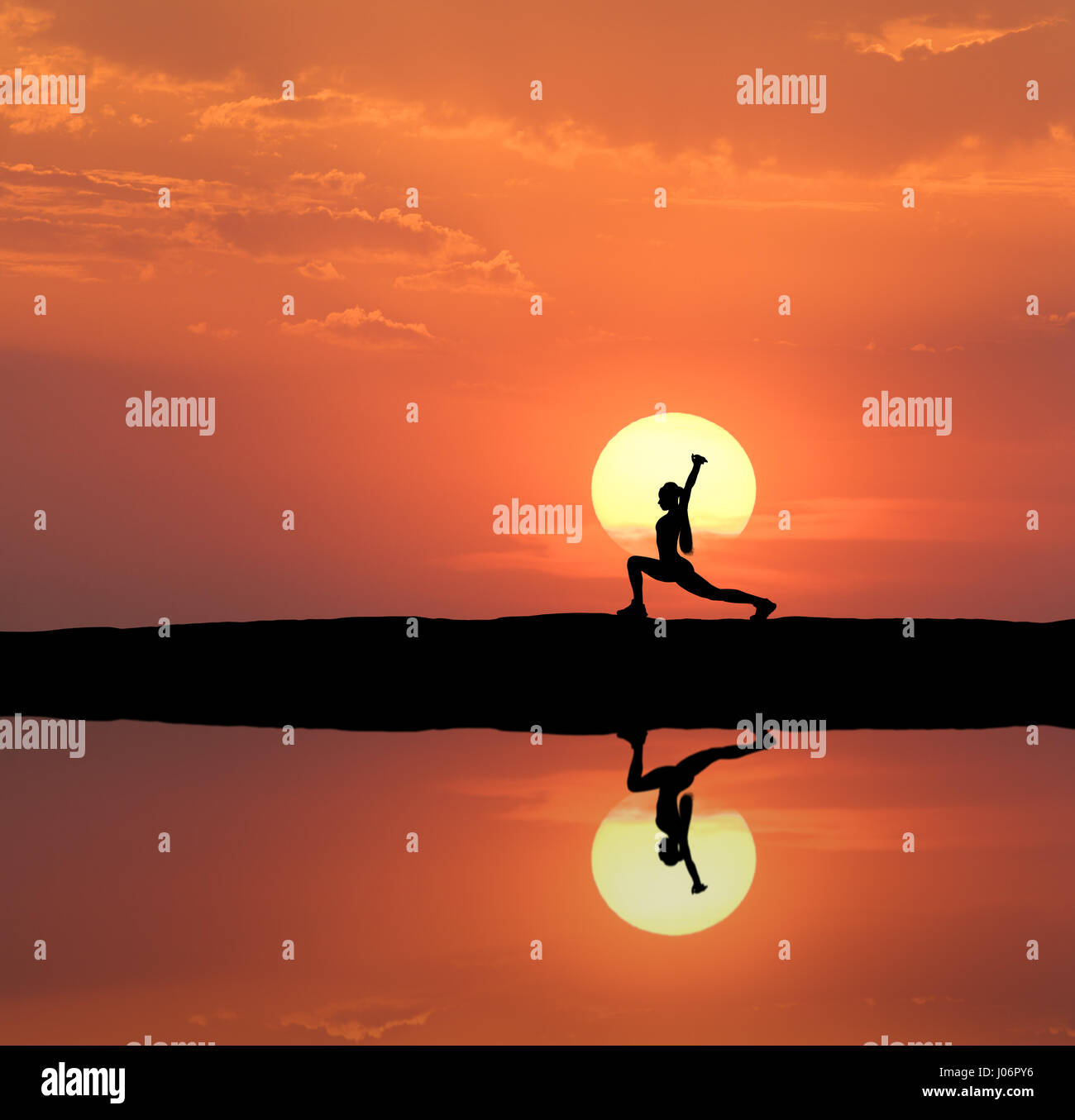 Silhouette of a standing sporty woman practicing yoga with raised up arms on the hill near the lake with sky reflection in water. Sun and orange sky w Stock Photo