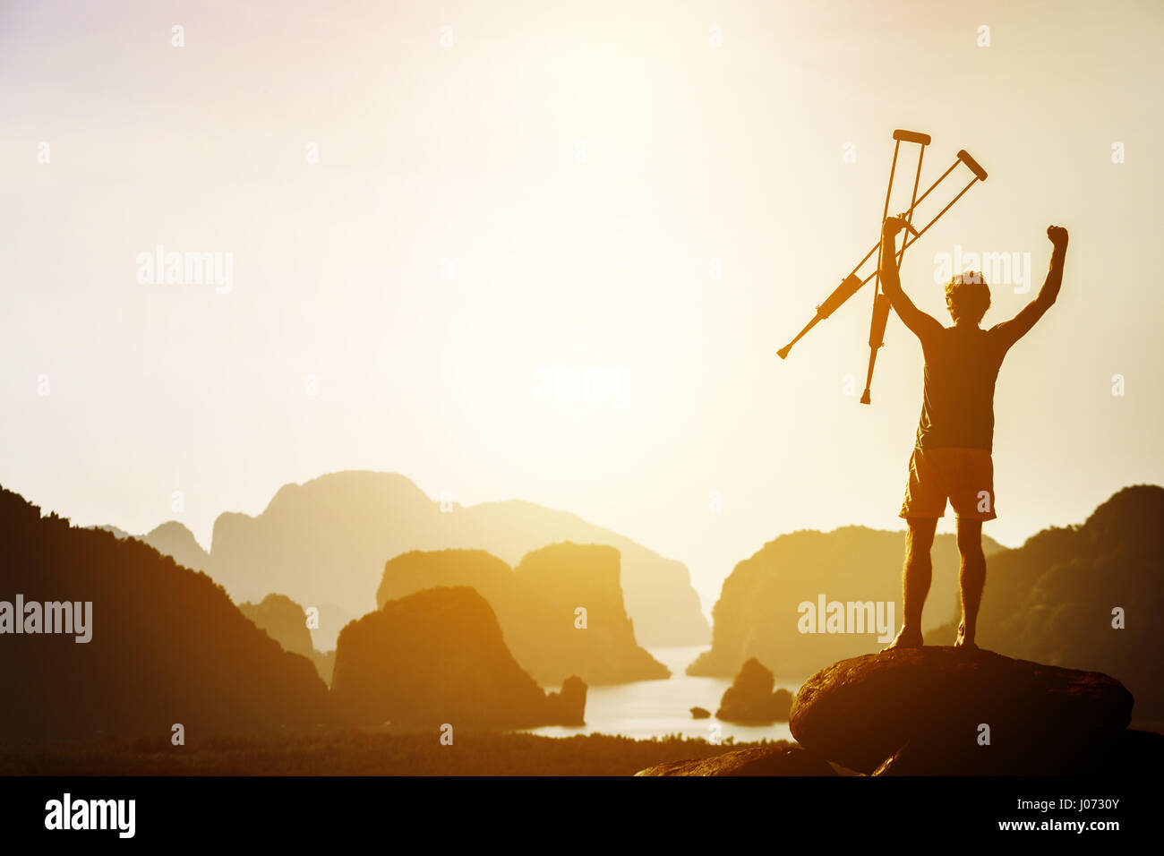 Man with crutches stands in winner pose with rised hands on mountains and islands background. Space for text Stock Photo