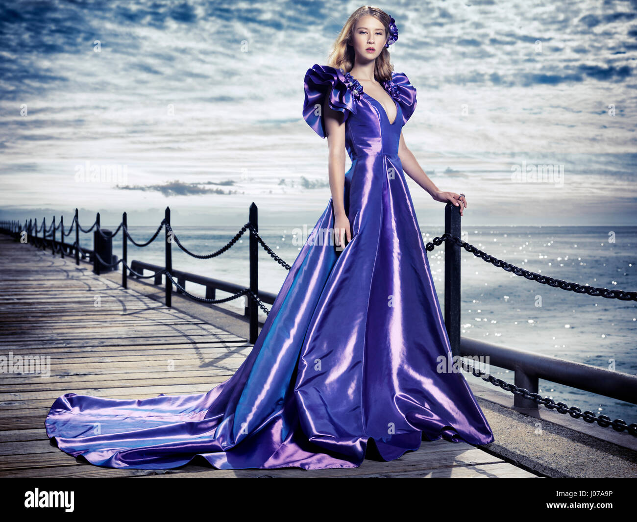 Young woman wearing a beautiful long blue evening gown standing at waterfront, artistic fashion portrait Stock Photo