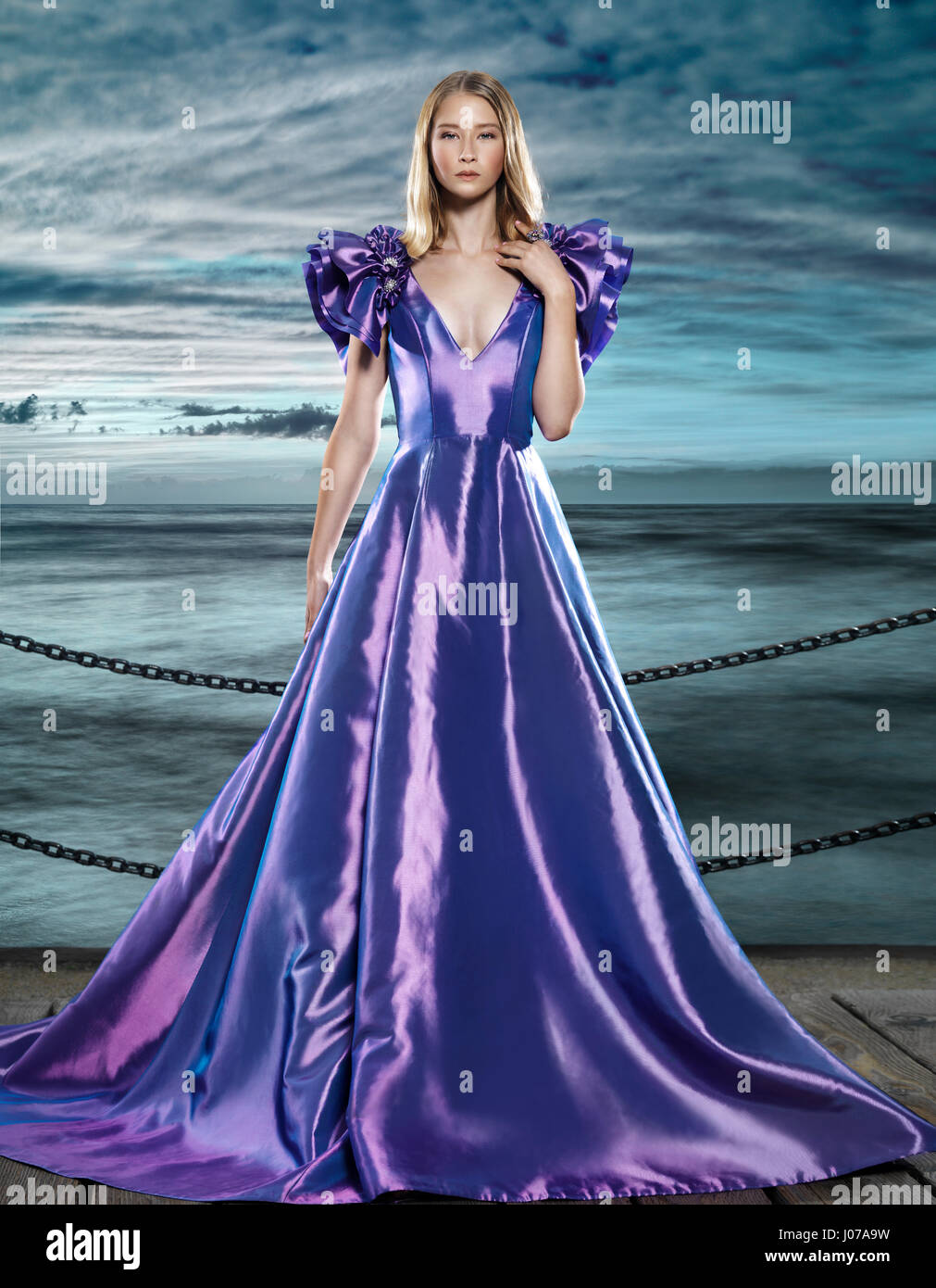 Young blond woman wearing a beautiful long blue dress, evening gown, at waterfront, artistic fashion portrait Stock Photo