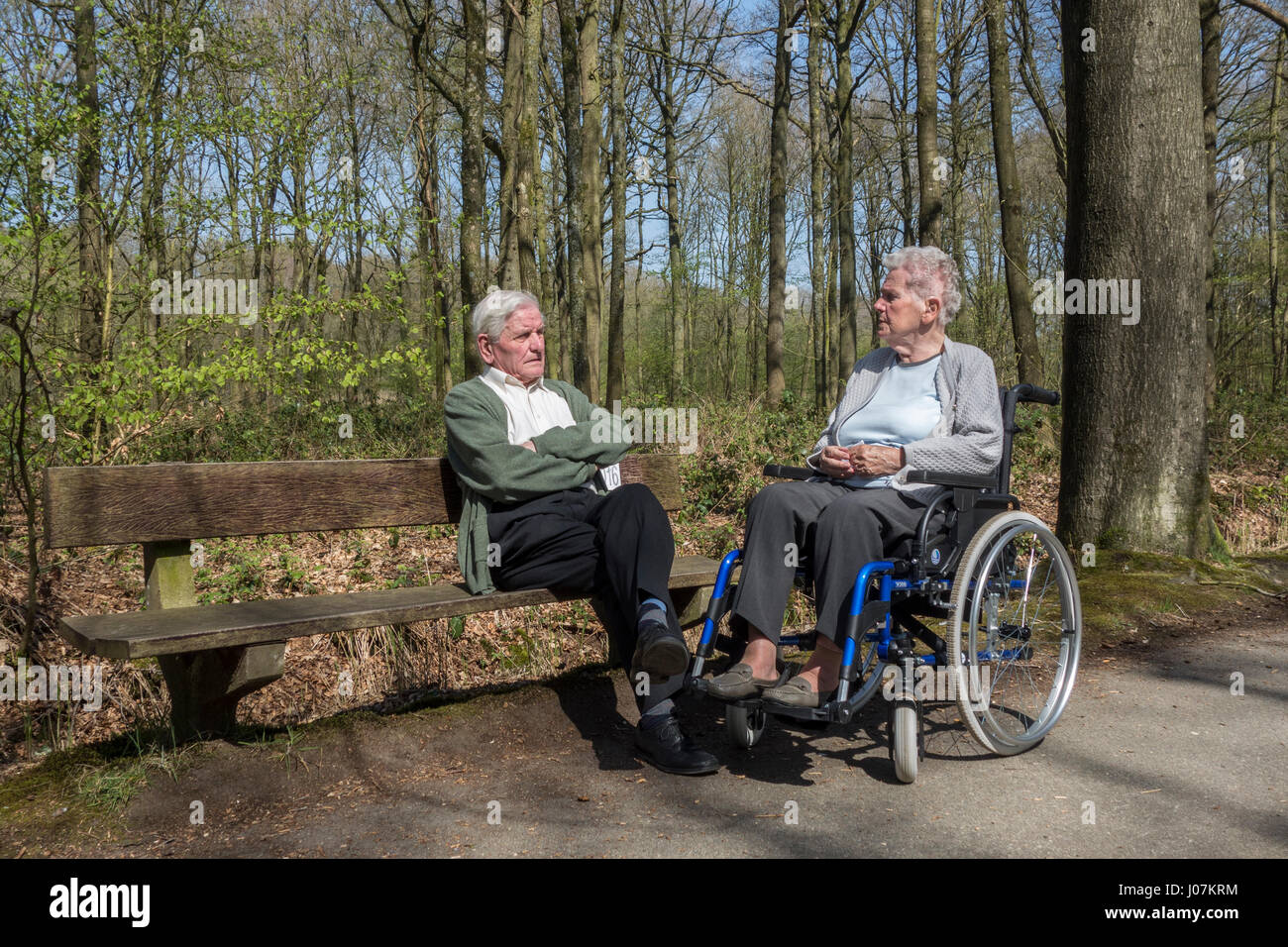 Disabled elderly woman in wheelchair talking to her retired husband sitting on a park bench during stroll in forest on a sunny day in spring Stock Photo