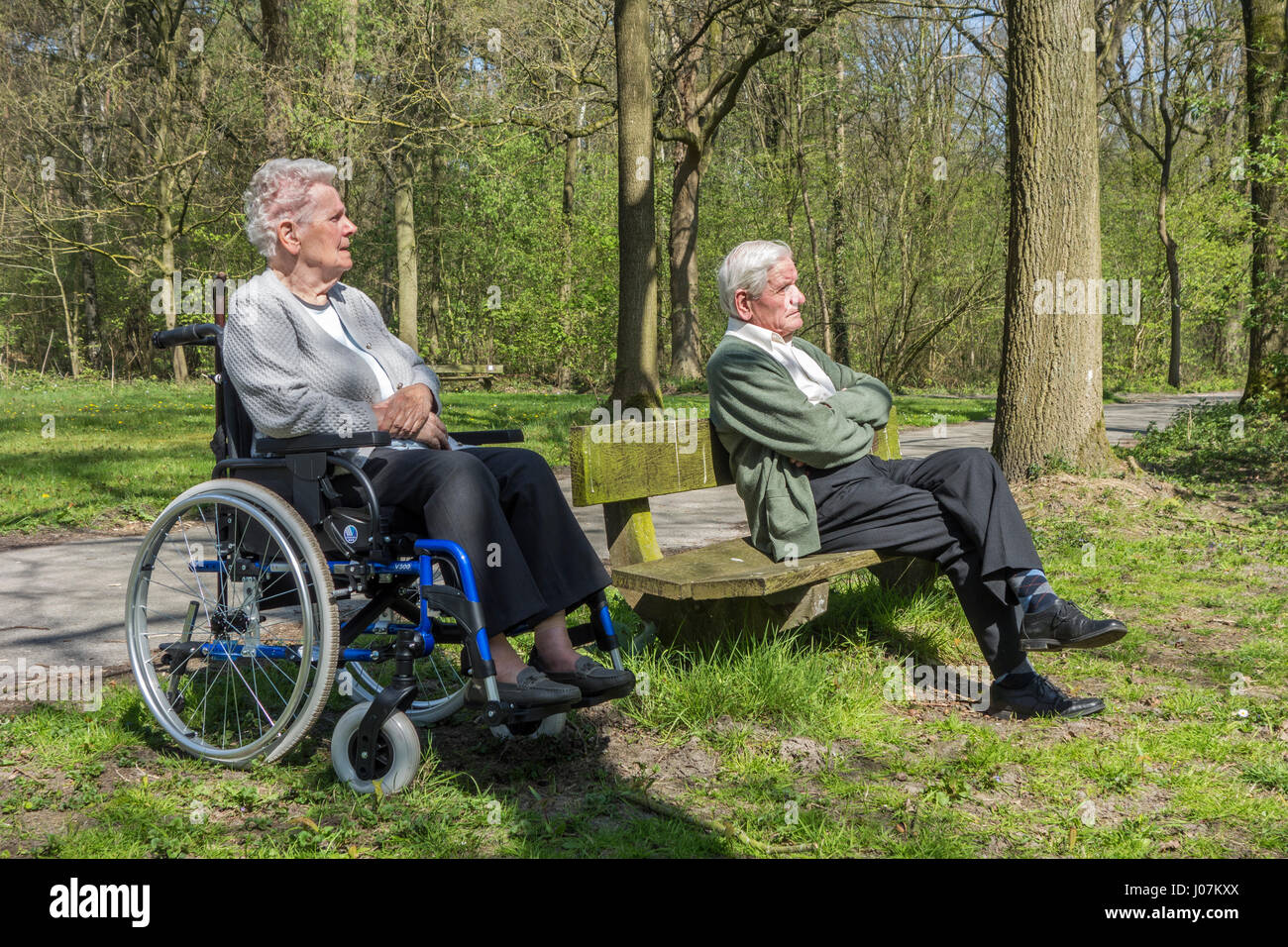 Disabled elderly woman in wheelchair and retired husband sitting on a park bench enjoying nature during stroll in forest on a sunny day in spring Stock Photo