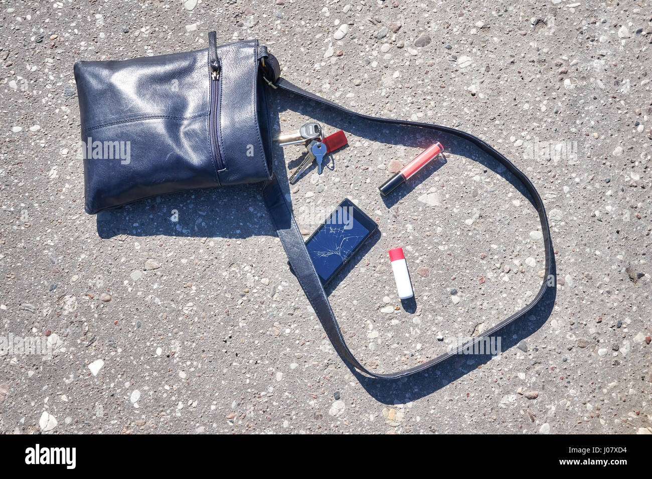 Handbag, cellular phone with broken screen, keys and lipstick on asphalt street, conceptual picture. Stock Photo