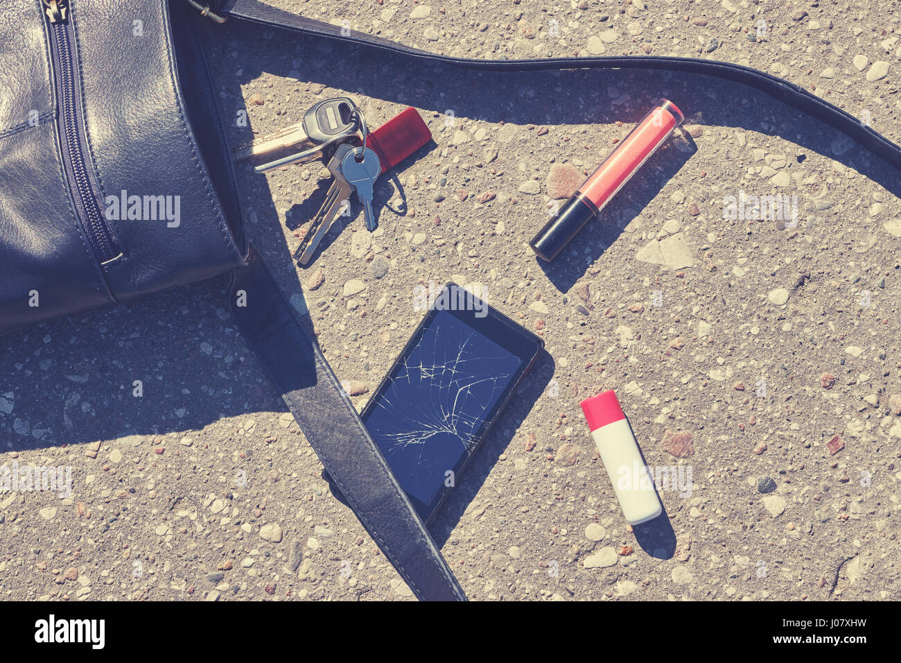 Close up of a handbag, cellular phone with broken screen, keys and lipstick on asphalt street, conceptual picture, color toning applied. Stock Photo
