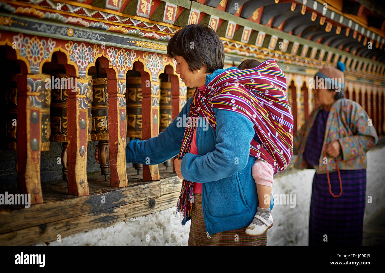 Two women, one carrying a baby, spin prayer wheels on the wall of the Changangkha Lhakhang Temple in Thimphu Bhutan Stock Photo