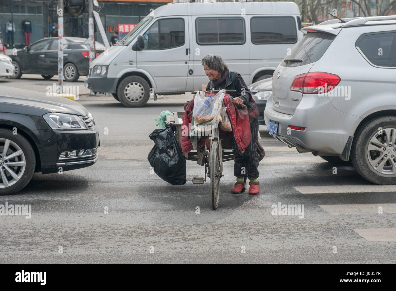An elderly poor woman pushes a tricycle through cars waiting traffic light in Xiong County, Hebei province, China. 09-Apr-2017 Stock Photo