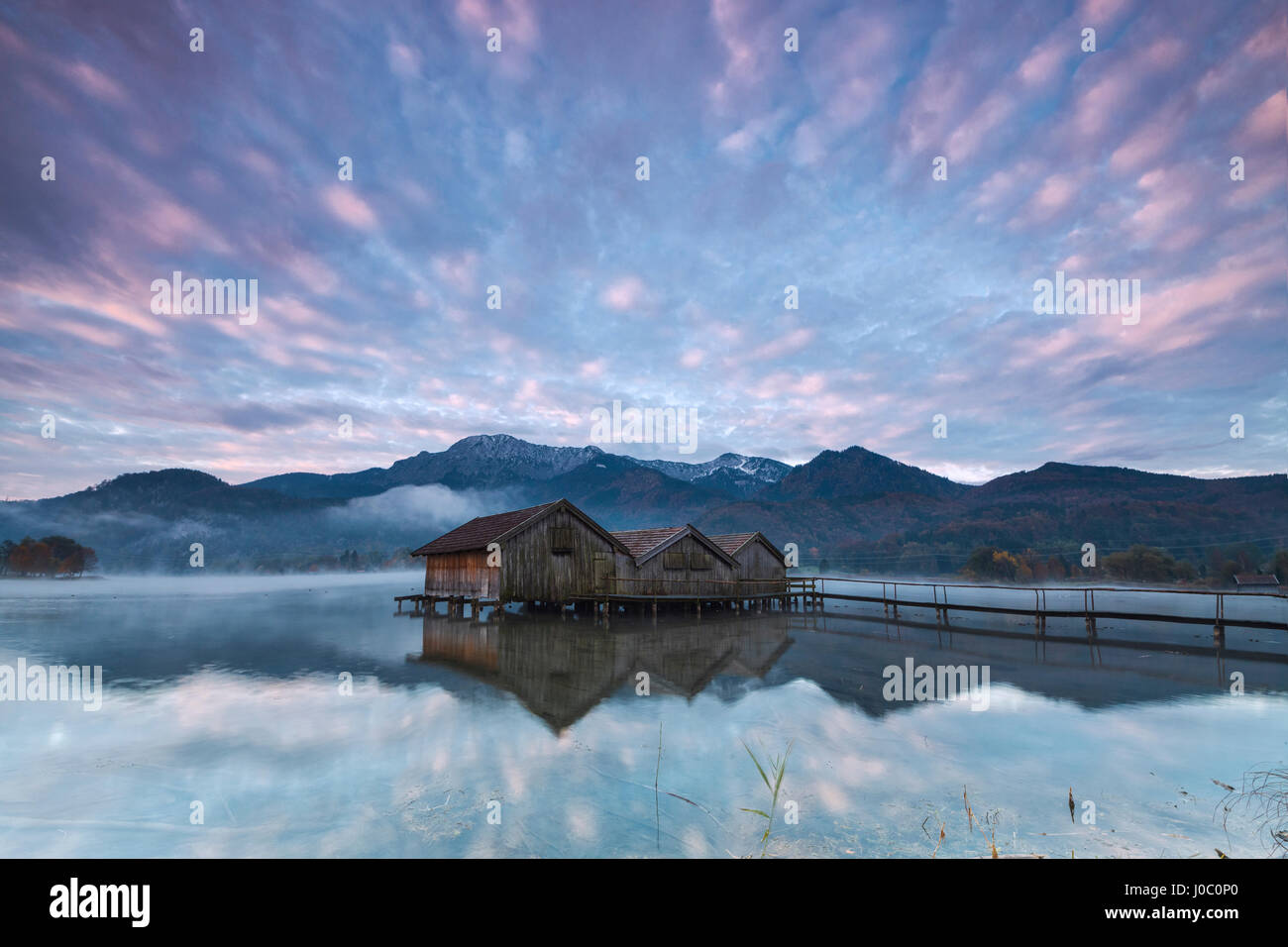 Pink clouds at sunset and wooden huts are reflected in the clear water of Kochelsee, Schlehdorf, Bavaria, Germany Stock Photo