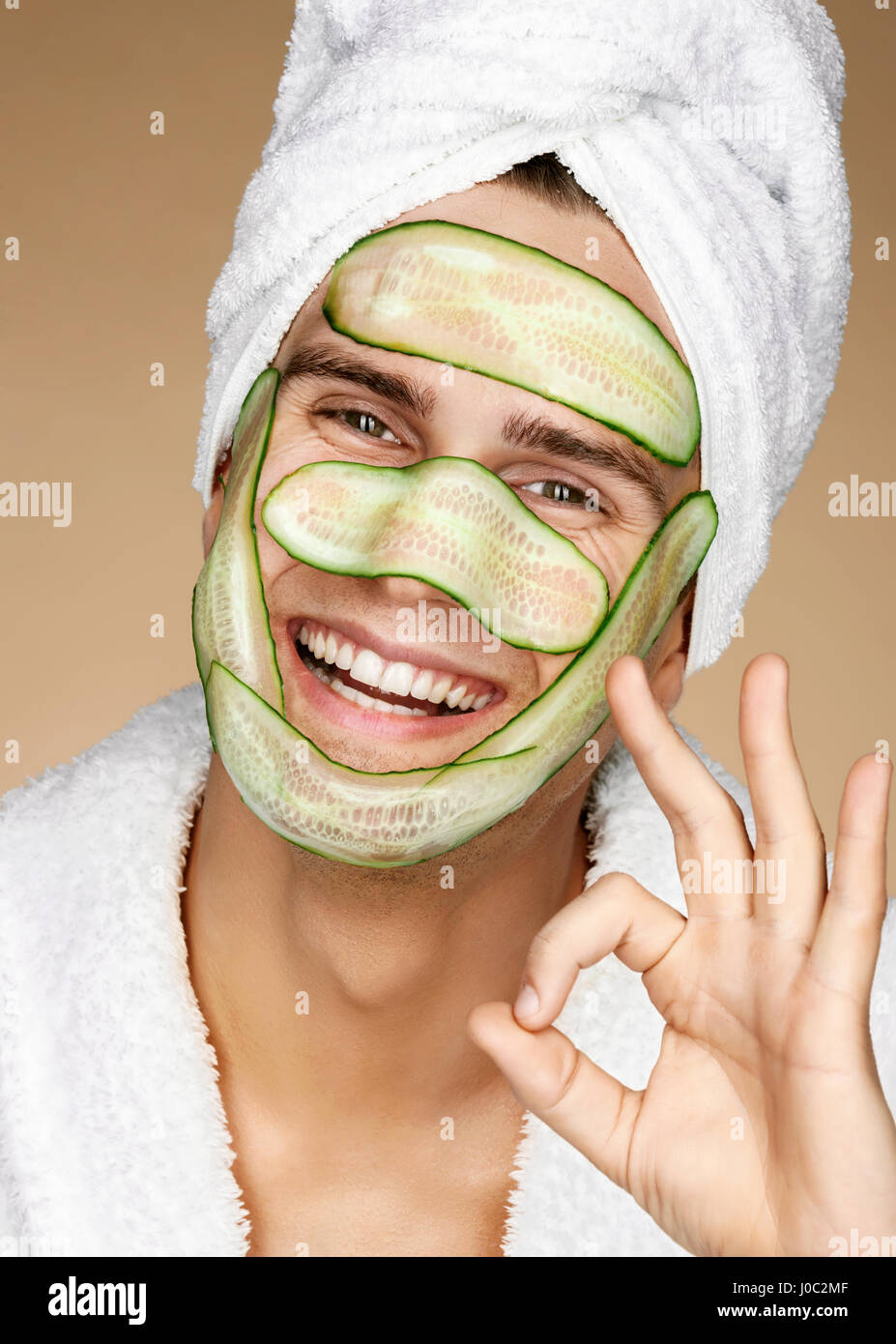 Funny man receiving facial mask of cucumber and showing okay gesture. Cosmetic procedure man's face. Grooming himself Stock Photo