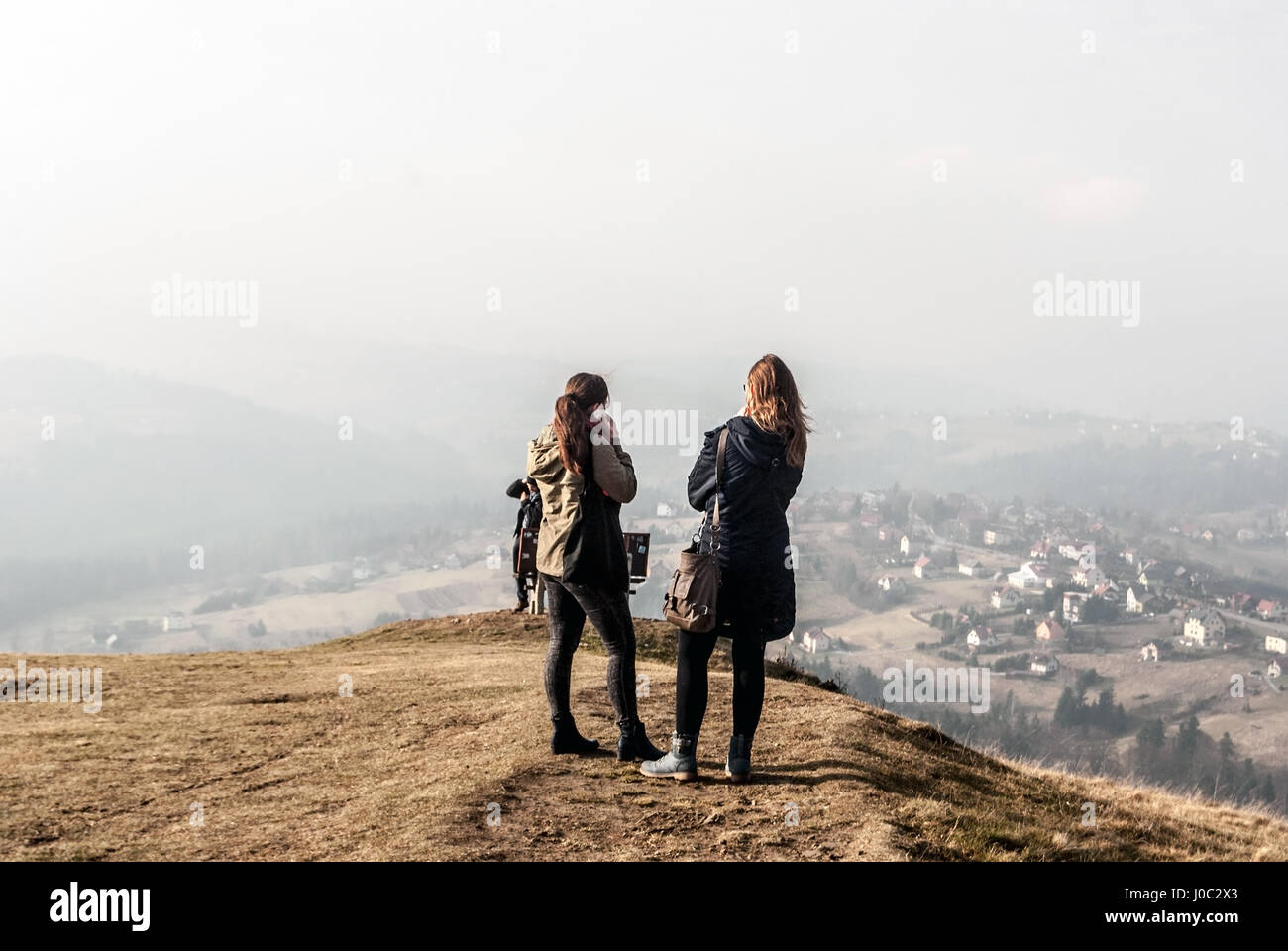 two young nice polish girls (one of them phoning) with long brown hair on Ochodzita hill summit with Koniakow village bellow in late autumn Silesian B Stock Photo