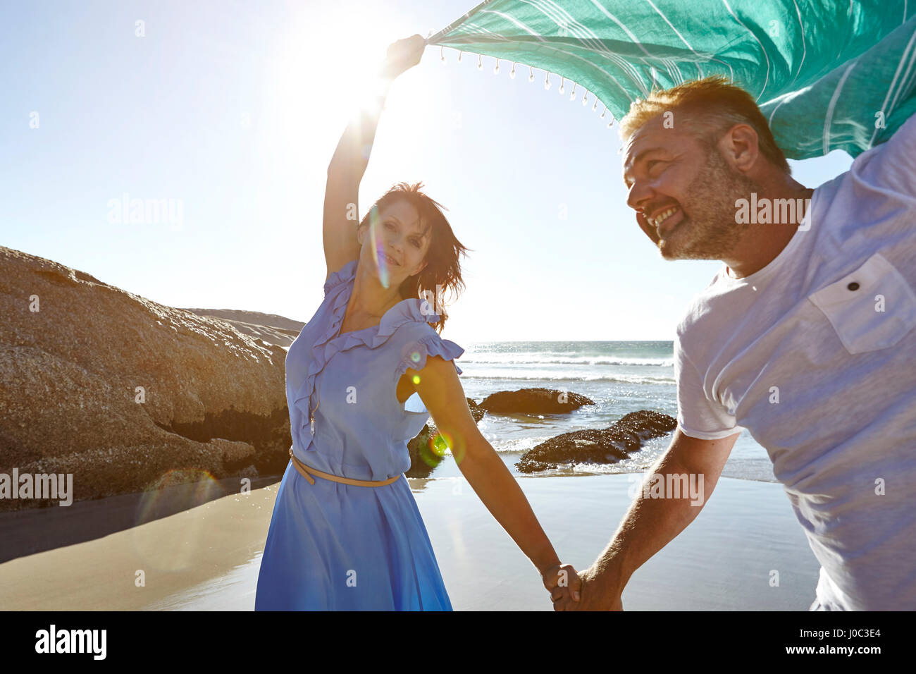 Mature couple holding hands on beach, woman holding sheer scarf in air Stock Photo