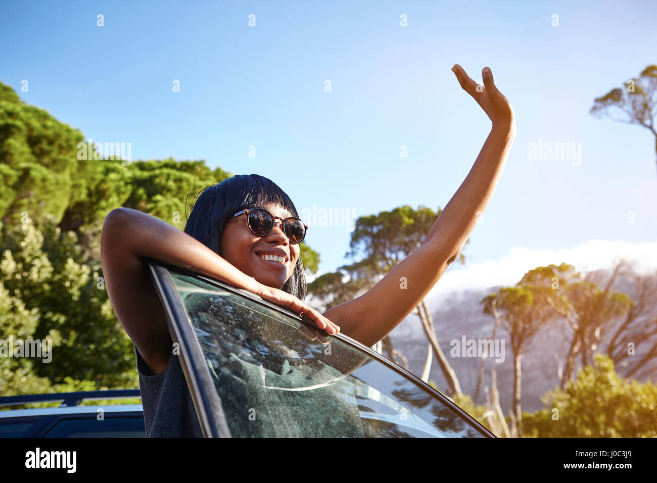 Portrait of young woman standing beside car, leaning on open car door, waving Stock Photo