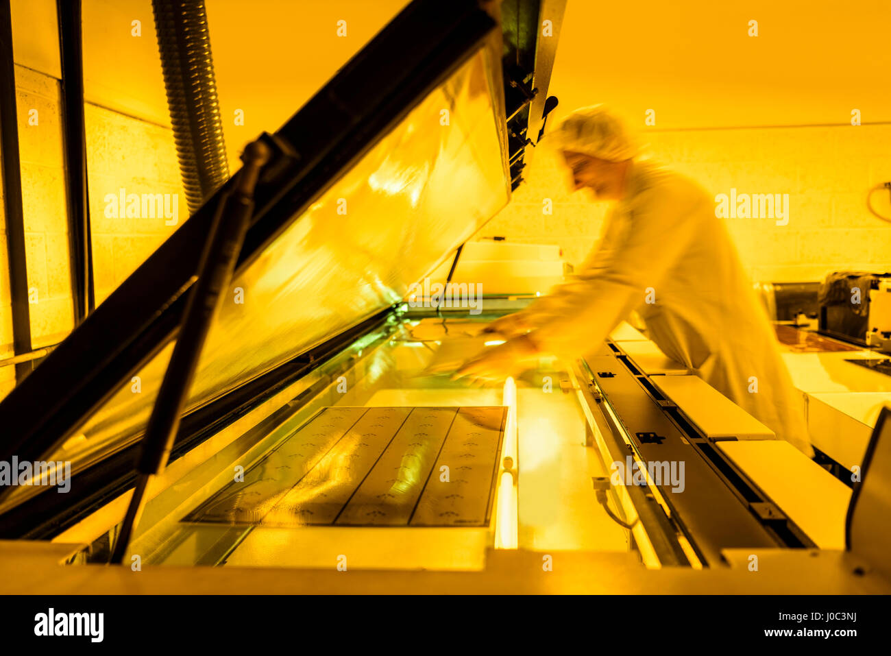 Worker placing negative of circuit board in machine in safe yellow light in electronics factory Stock Photo