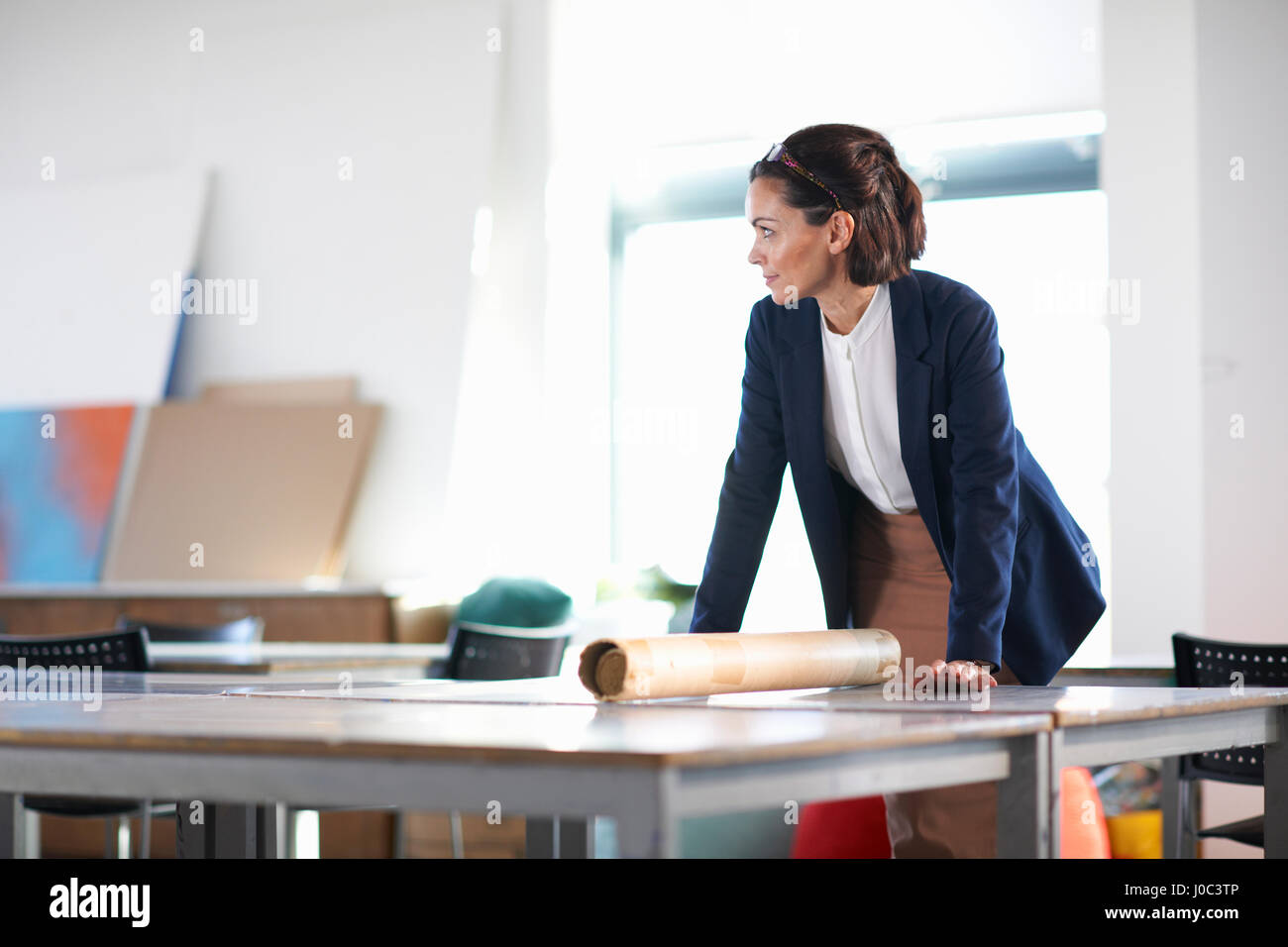Woman in office leaning against desk looking away Stock Photo - Alamy