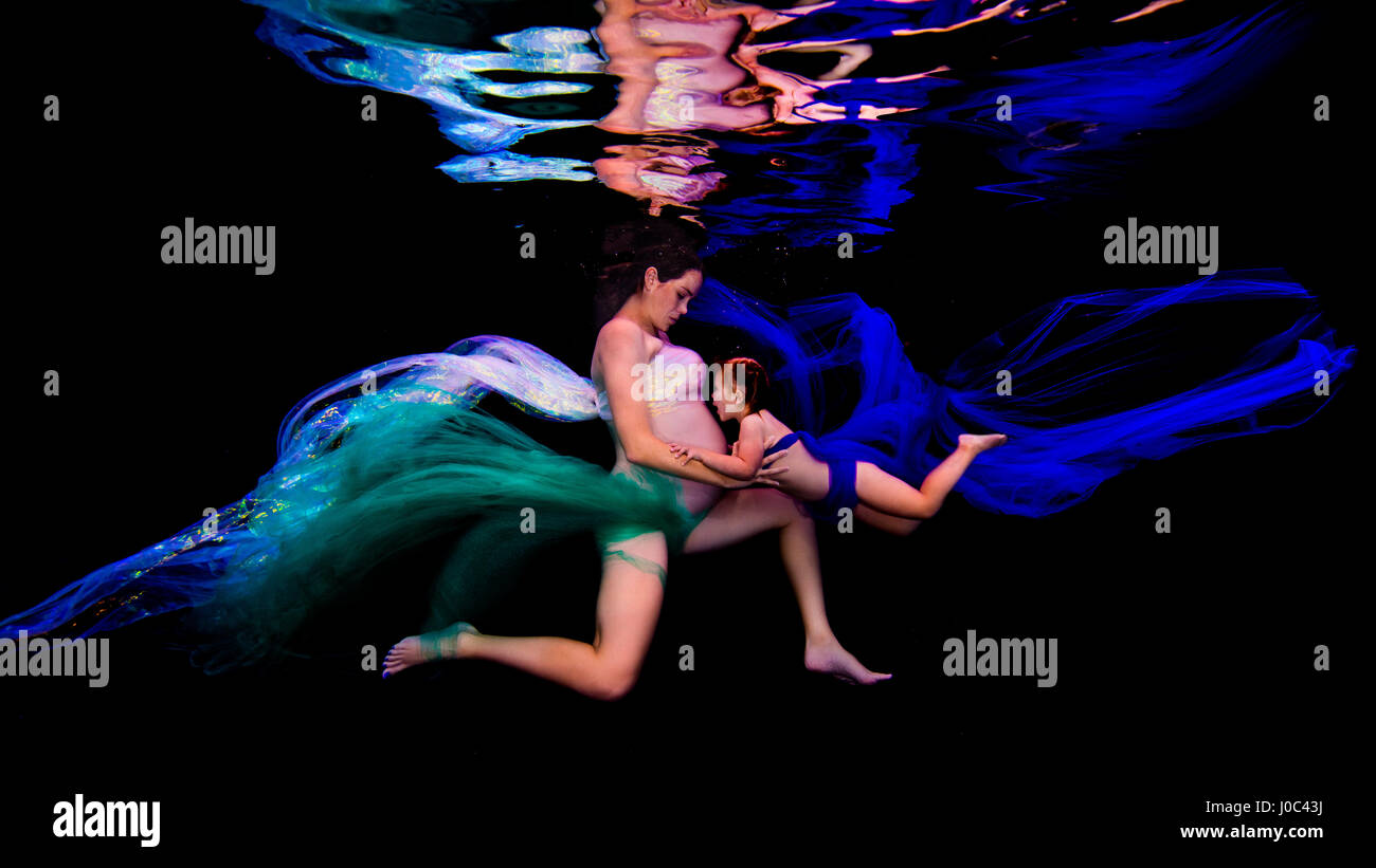 Underwater view of young pregnant woman and girl poised together in darkness Stock Photo