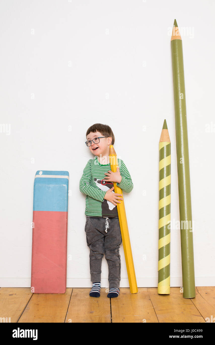 Young boy standing, holding giant size pencil, giant stationery leaning on wall beside him Stock Photo