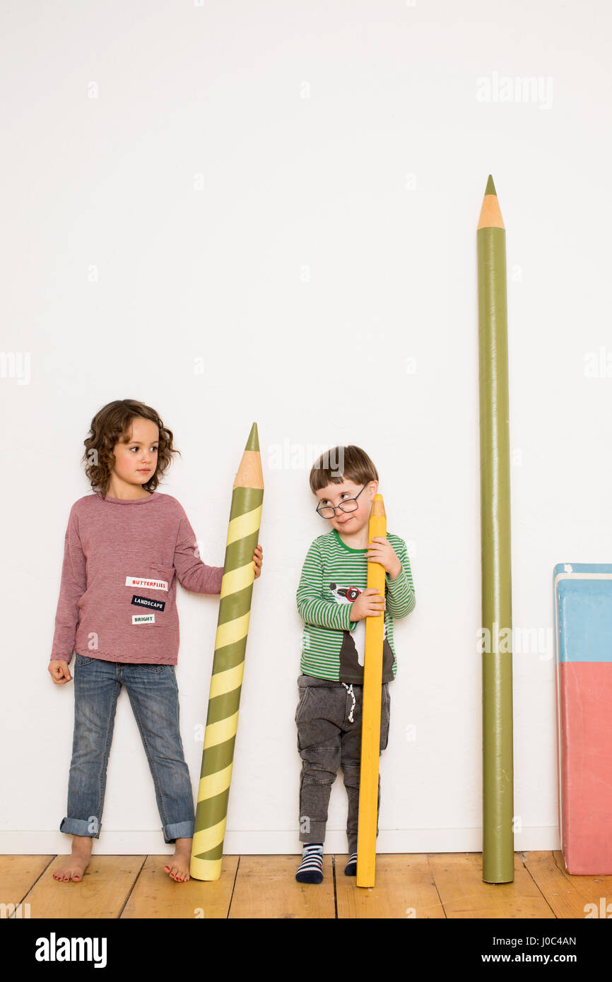 Young girl and boy standing, holding giant size pencils, giant stationery leaning on wall beside him Stock Photo