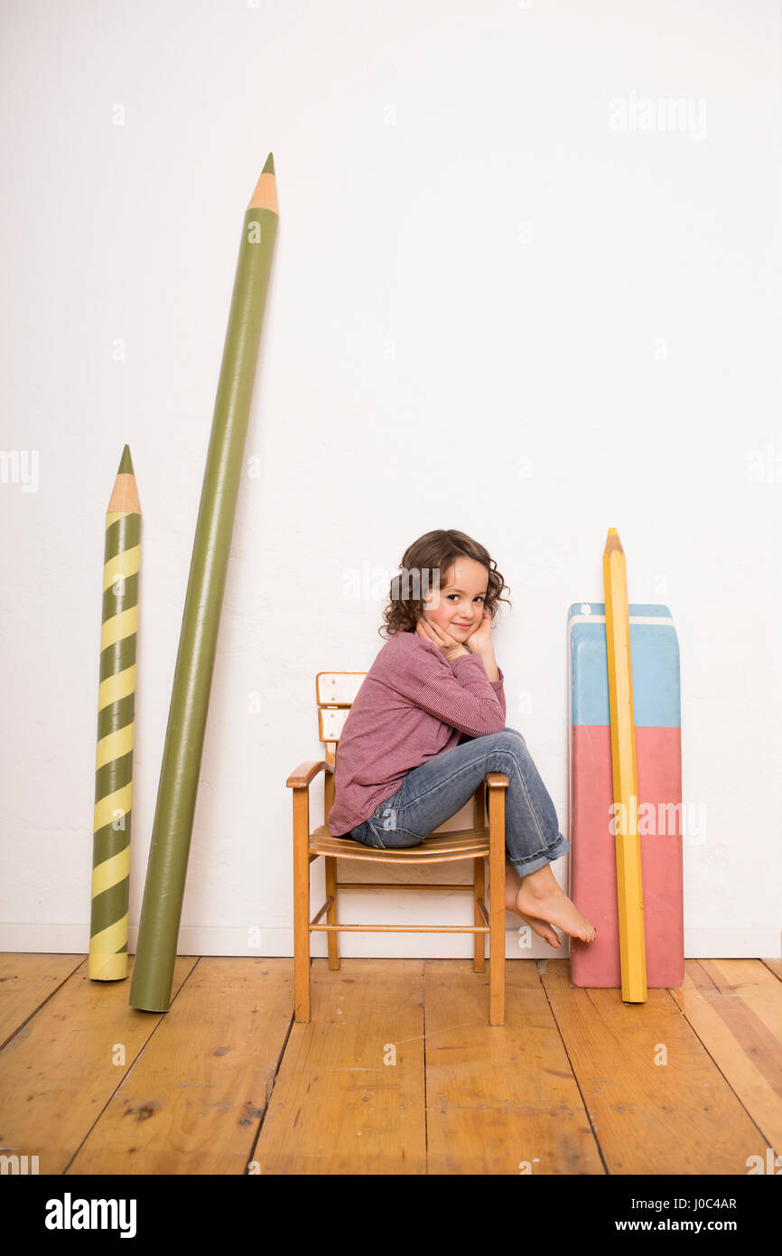 Young girl sitting on chair, giant size stationery leaning on wall beside her Stock Photo