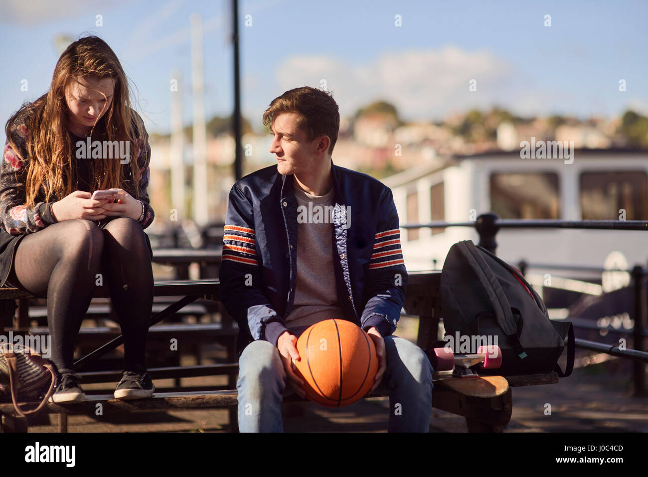 Two friends sitting outdoors, young man holding basketball, young woman using smartphone, Bristol, UK Stock Photo