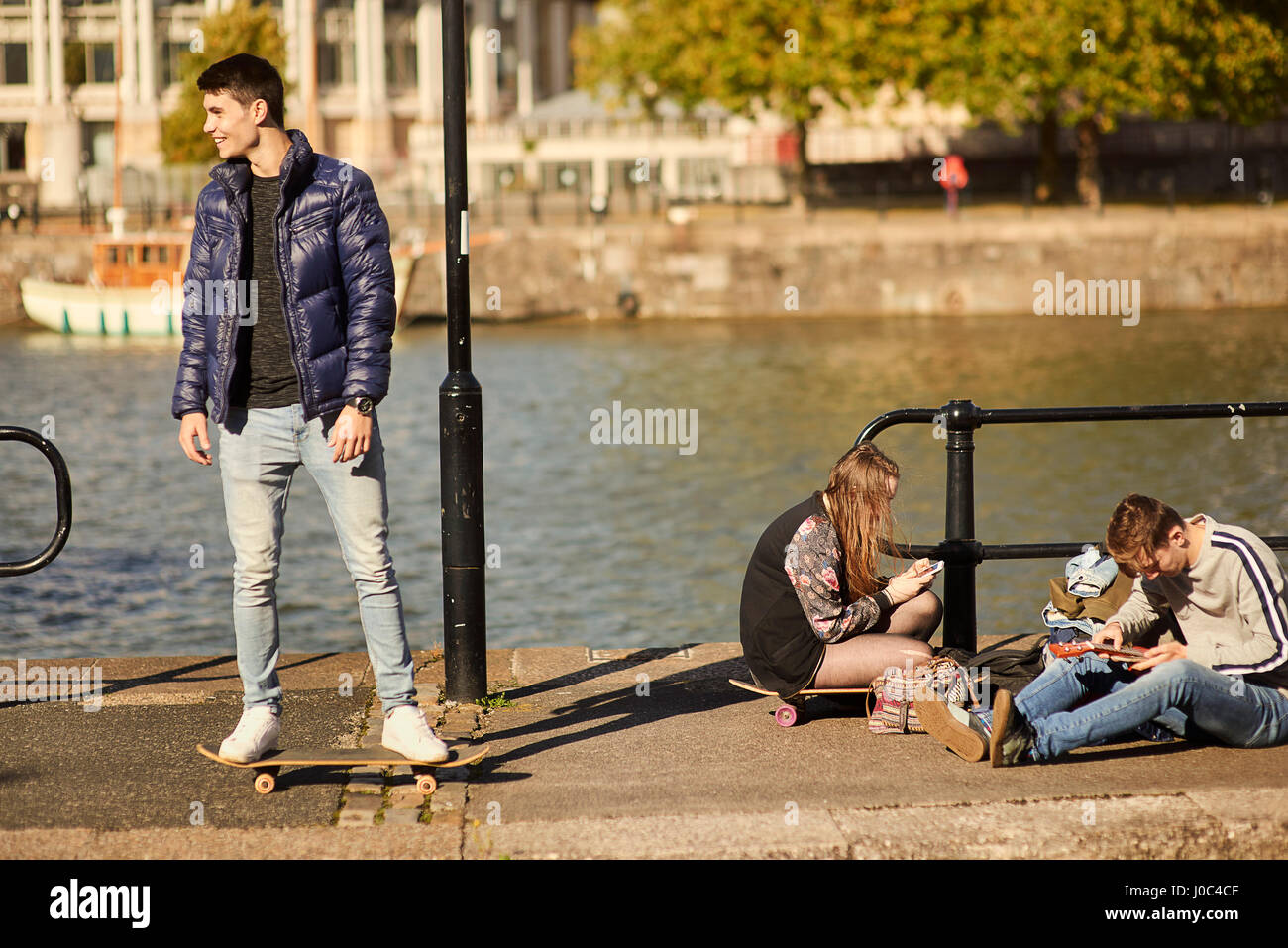 Three friends fooling around beside river, young man on skateboard, Bristol, UK Stock Photo