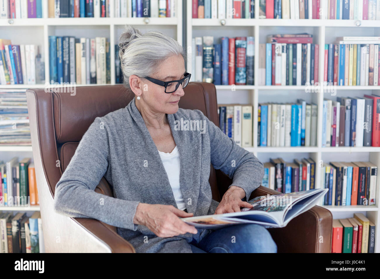 Senior woman sitting in chair in library, reading book Stock Photo
