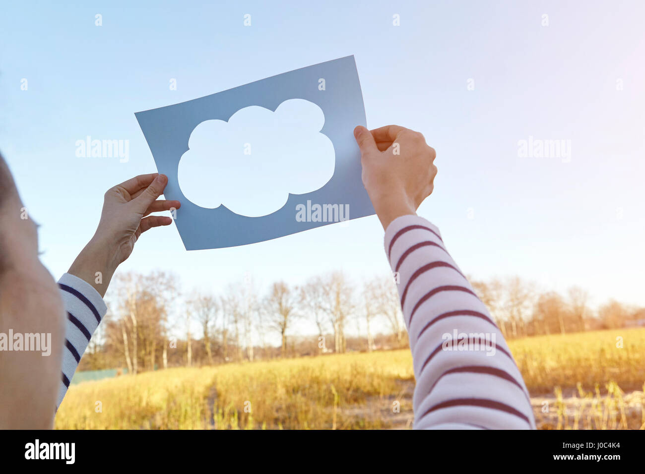 Woman standing in rural setting, holding paper with cut-out of cloud, towards sky Stock Photo