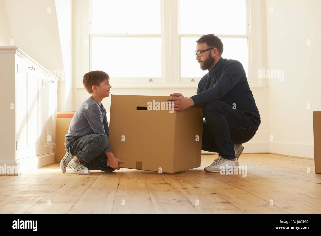 Mid adult man and son crouching to lift cardboard box in new home Stock Photo