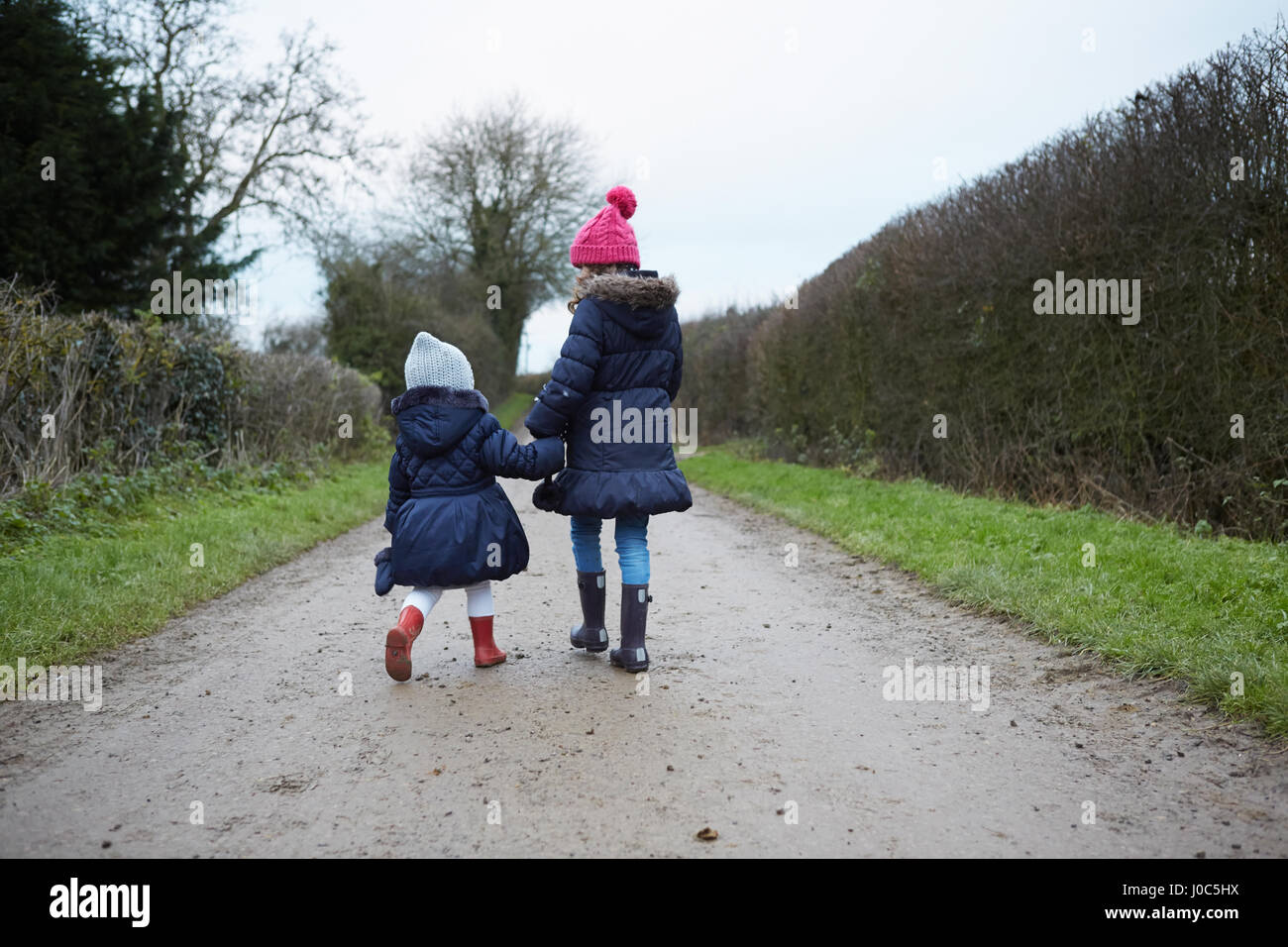 Rear view of girl and female toddler in knit hats walking along rural road Stock Photo
