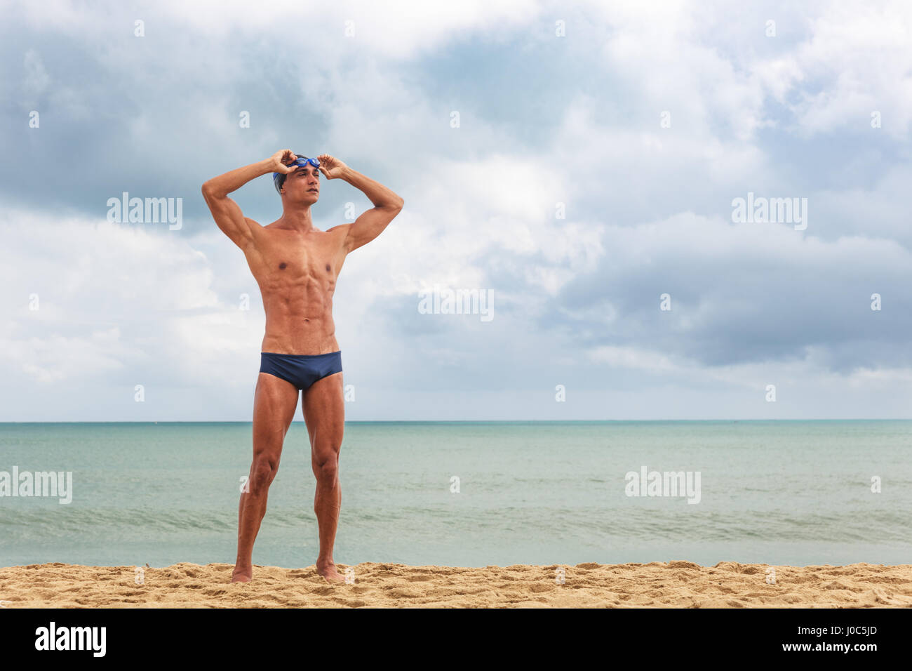 Muscular male swimmer standing on beach putting on swimming goggles Stock Photo