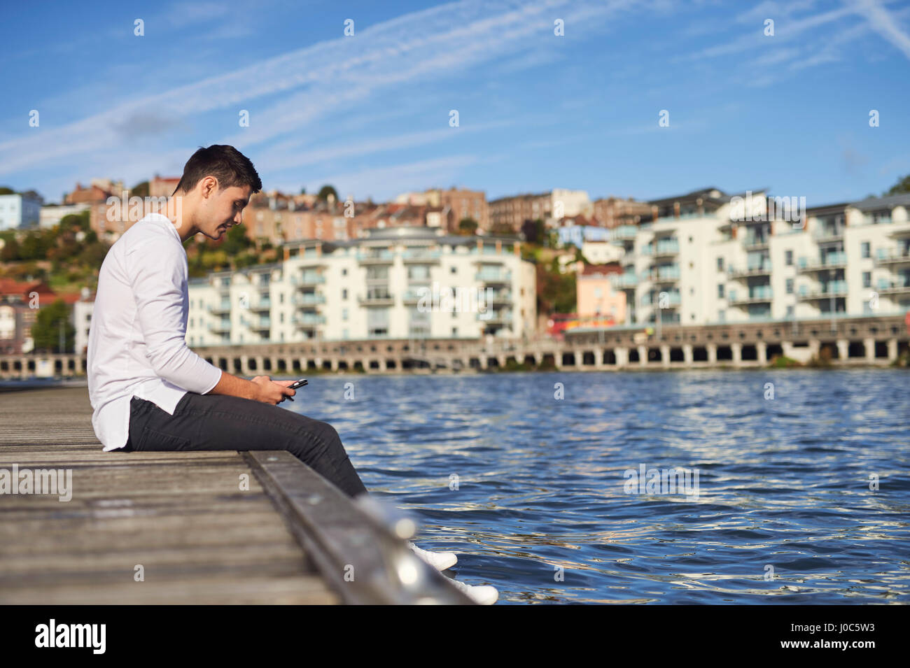 Young man sitting by river, using smartphone, Bristol, UK Stock Photo