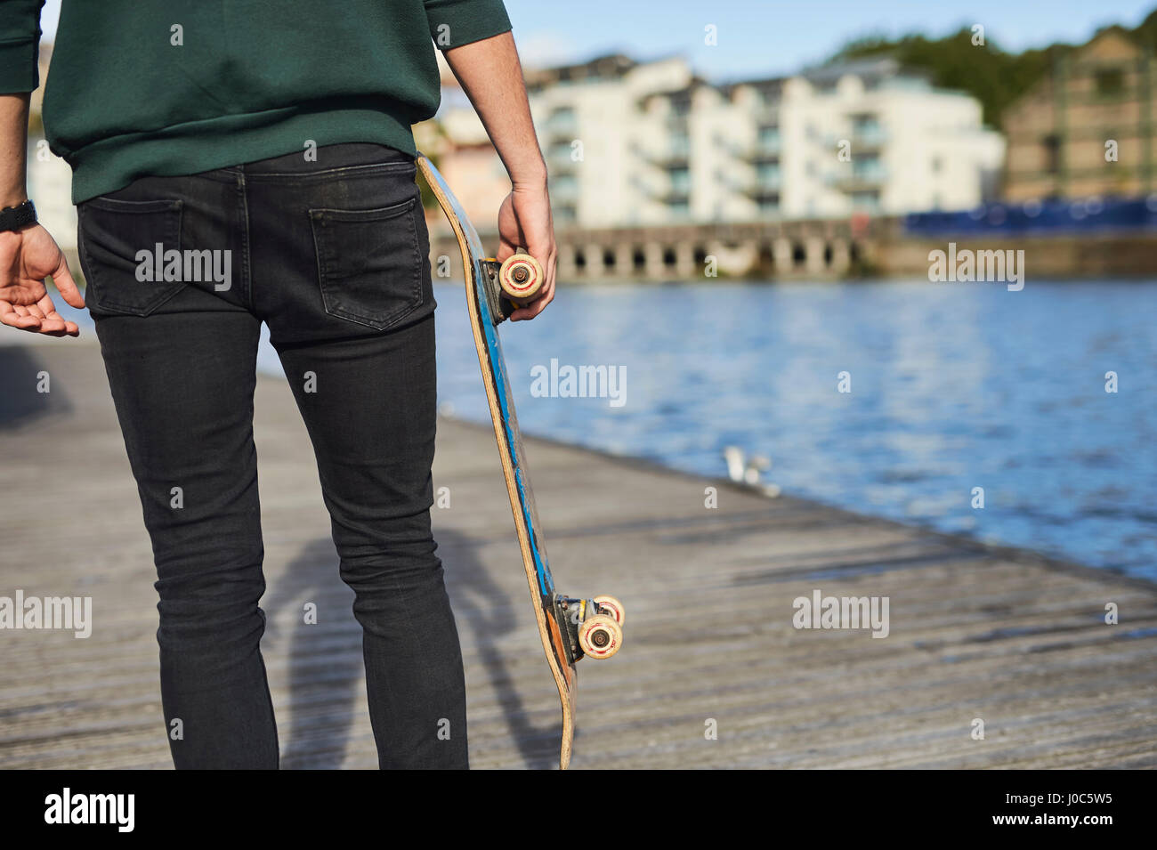 Young man walking beside river, holding skateboard, rear view, mid section, Bristol, UK Stock Photo
