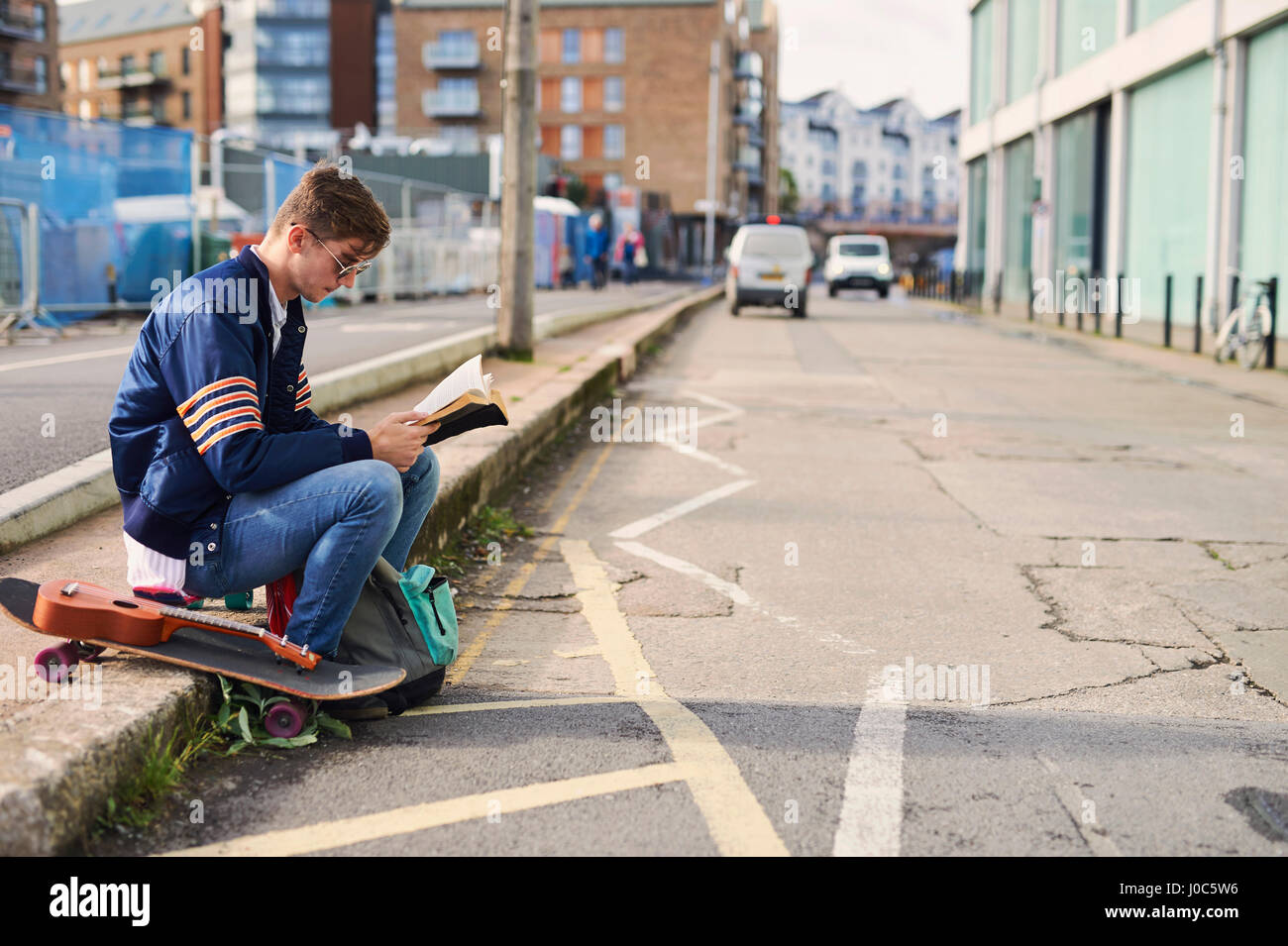 Young man sitting on kerb, reading book, skateboard beside him, Bristol, UK Stock Photo