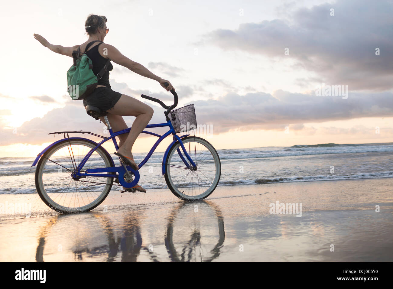 Woman waving while cycling on beach at sunset, Nosara, Guanacaste Province, Costa Rica Stock Photo