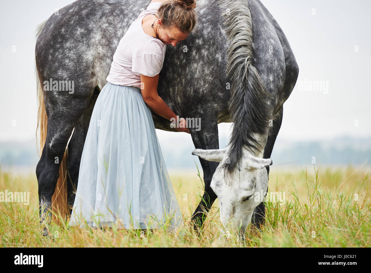 Woman in skirt with arms around dapple grey horse in field Stock Photo