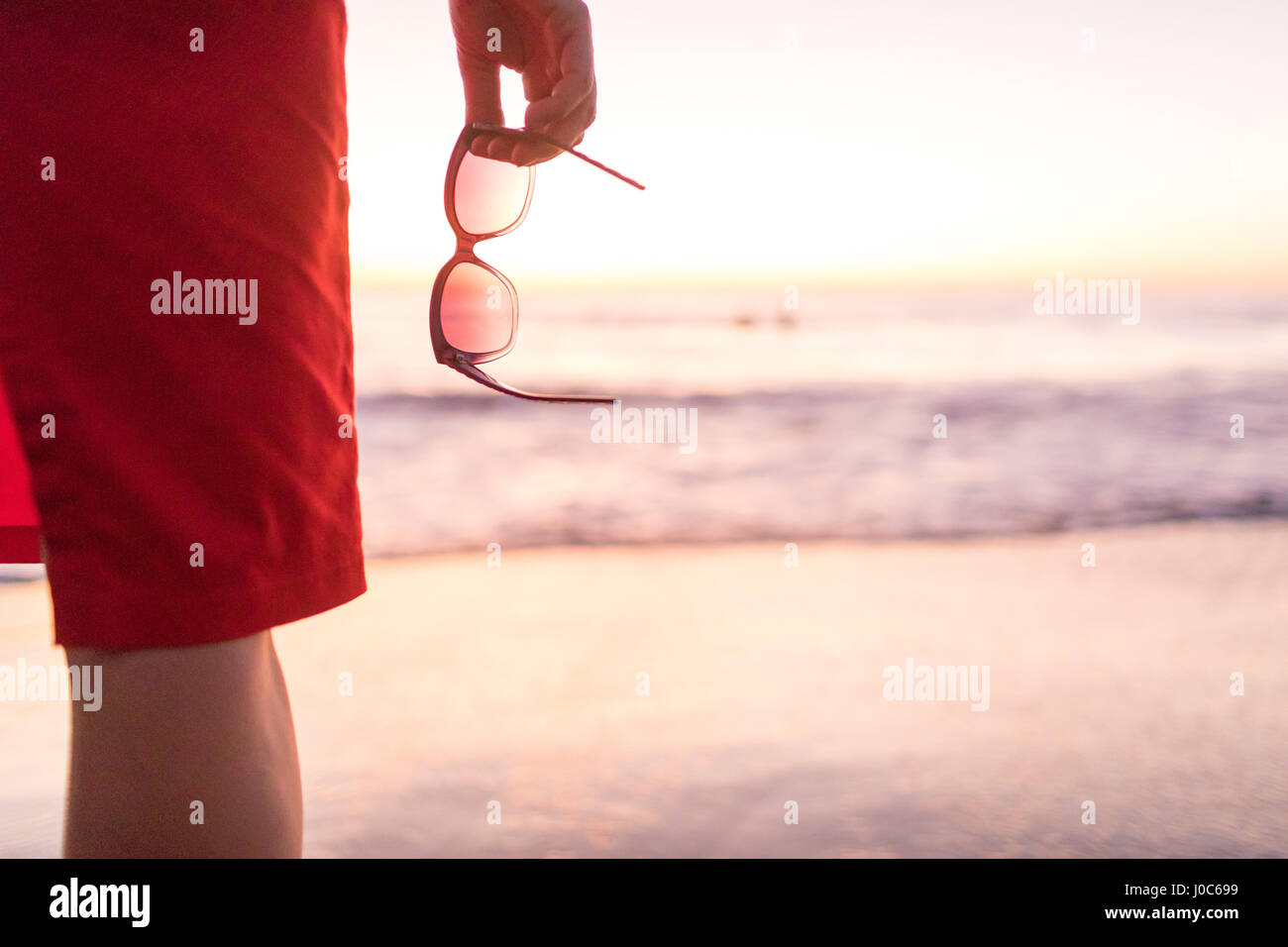 Rear cropped view of woman's hand holding sunglasses on beach at sunset, Nosara, Guanacaste Province, Costa Rica Stock Photo