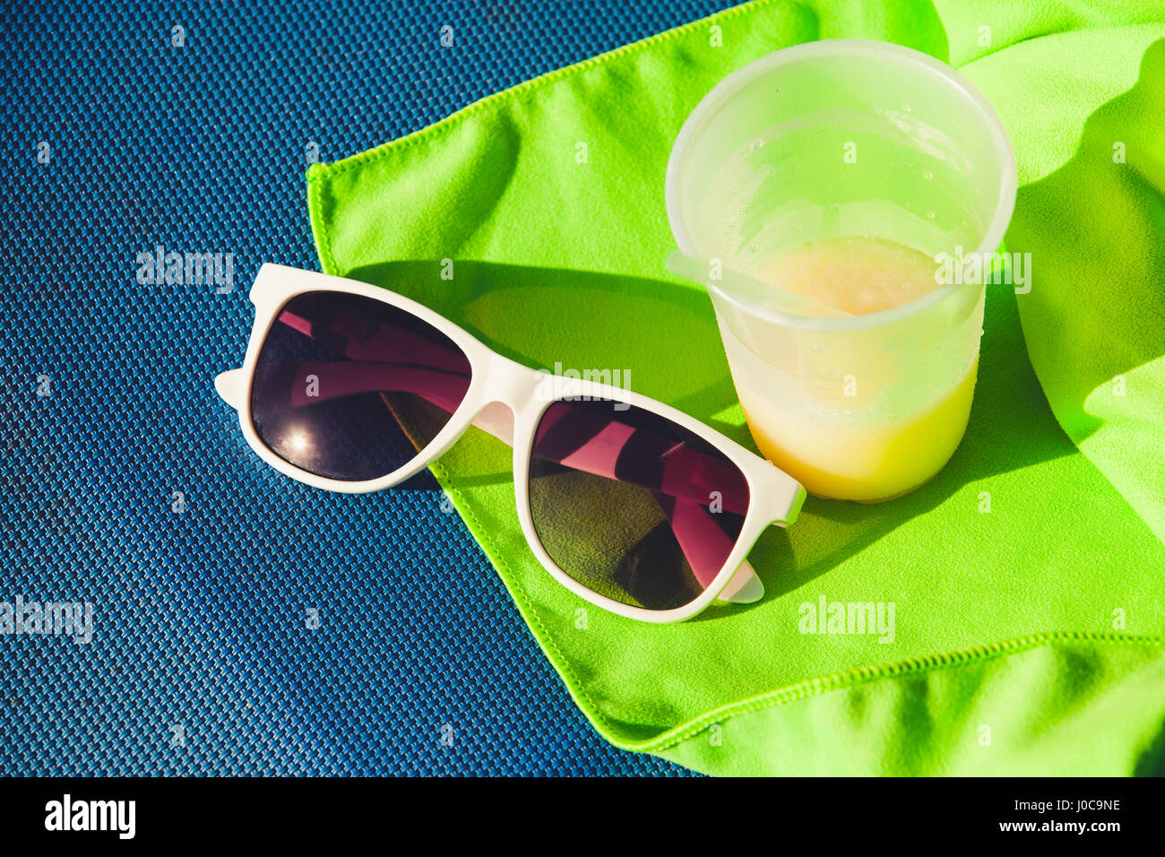 Sunglasses and plastic glass of juice stand on green towel over blue sun lounger, summer beach resort vacation theme Stock Photo